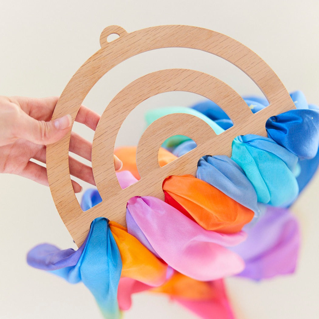 A hand holds a wooden Rainbow Playsilk Display, with colorful silk scarves threaded through it—perfect for brightening up any playroom.
